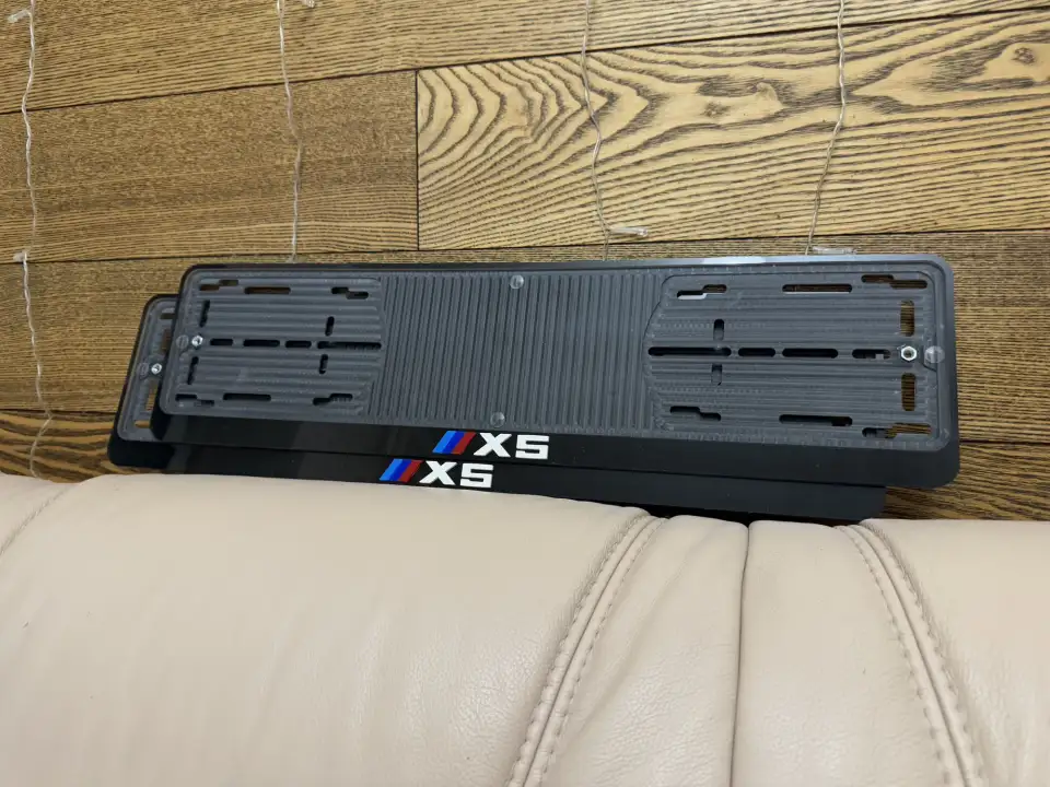 BMW X5 · чёрные рамки с M-полосками Две чёрные номерные рамки BMW X5 с эмблемой M на деревянной стене