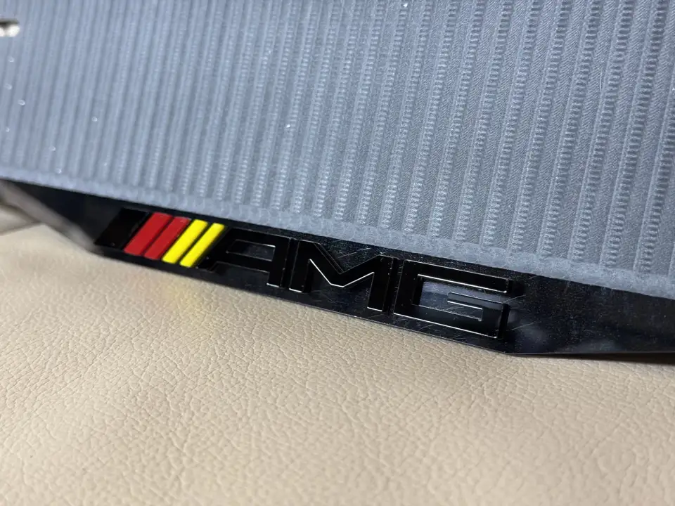 Mercedes-AMG · логотип крупным планом Макро объёмного логотипа AMG с красно-жёлтыми полосами на чёрной глянцевой номерной рамке Mercedes-AMG