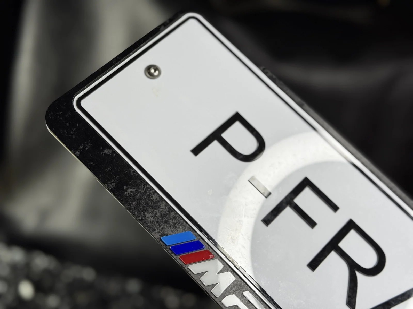 Квадратная рамка для номера в цвете карбон с логотипом M: белые буквы и голубая, синяя и красная полосы на планке