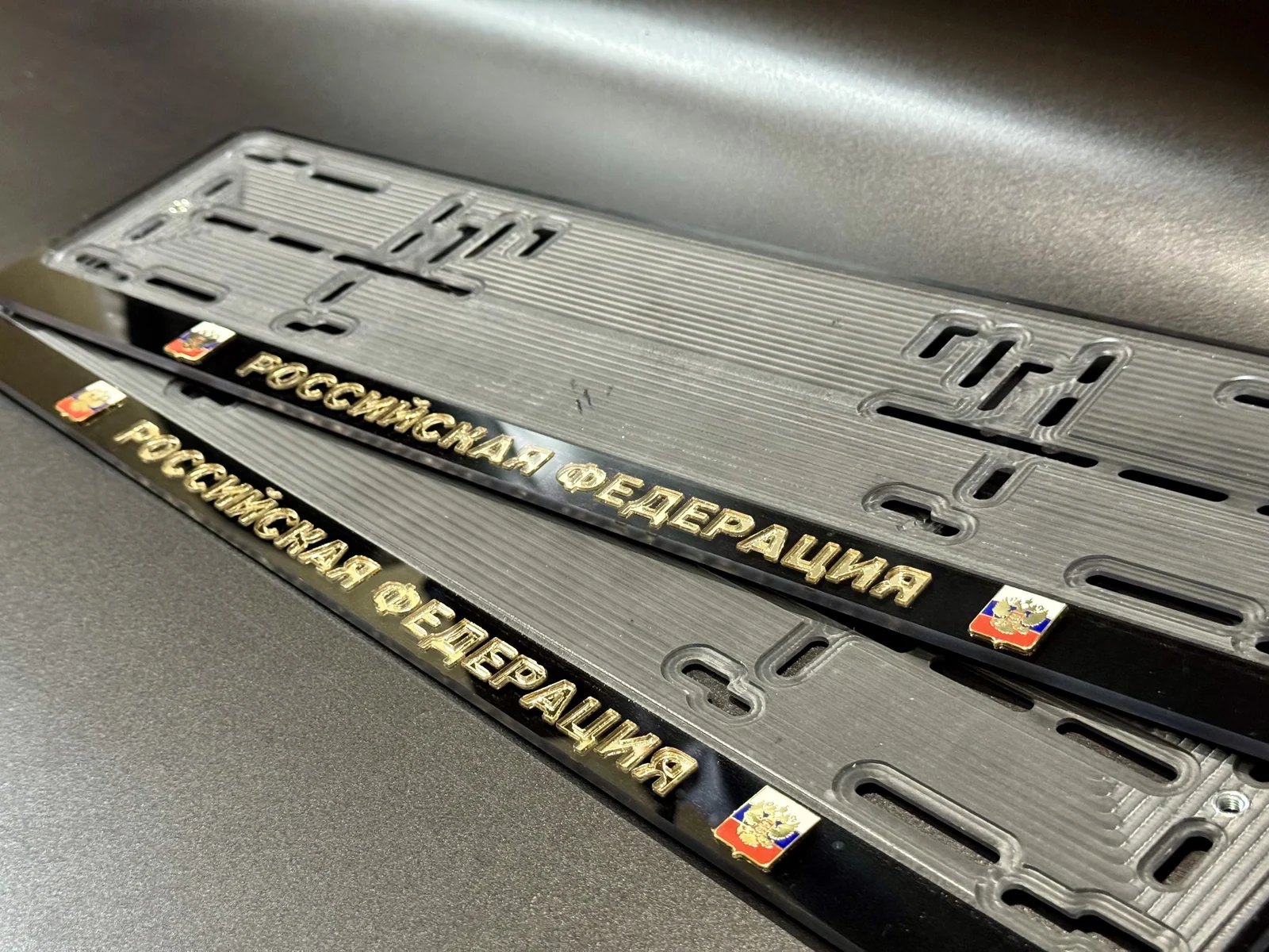 Две рамки для номера: чёрный глянец, золотые буквы РОССИЙСКАЯ ФЕДЕРАЦИЯ и значки с гербом на фоне триколора по краям планки